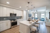 Located at 5050 Aspire Way in Naples, FL, this well-appointed 2-bedroom, 3-bathroom residence offers a thoughtfully designed single-floor layout with a clean, coastal-inspired aesthetic. Light wood flooring flows throughout the main living areas, complementing the bright, neutral palette that defines the space.

The open-concept kitchen and living room serve as the heart of the home. The kitchen features white shaker cabinetry with gold hardware, a light blue subway tile backsplash, stainless steel appliances, and a central island with a quartz countertop and bar seating illuminated by pendant lighting. The living area is generously sized, with large windows and a private balcony access that brings in abundant natural light and outdoor views.

The spacious primary bedroom includes plush carpeting, a ceiling fan, and direct access to a well-sized closet and en-suite bathroom. The second bedroom is equally comfortable with similar finishes. The bathrooms are finished in crisp white tones — one featuring a dual-sink vanity with gold hardware and a glass-enclosed walk-in shower, and another with a full soaking tub and shower combination. Additional highlights include a dedicated laundry area, a utility room, a hallway, and three closets, ensuring ample storage throughout.