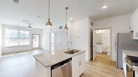 This well-designed single-floor residence offers a clean, contemporary aesthetic throughout, characterized by crisp white walls, light wood-style flooring, and an abundance of natural light. The open-concept kitchen serves as the heart of the space, anchored by a central island with granite countertops, an undermount sink, and stylish pendant lighting overhead. White shaker cabinetry lines the walls, complemented by a full suite of stainless steel appliances including a refrigerator, electric range, over-the-range microwave, and dishwasher — offering a well-equipped cooking environment. The kitchen flows naturally into the living area, which features a ceiling fan, large windows, and direct access to an exterior door, creating a bright and airy atmosphere. The bedrooms are generously sized, with plush carpeting, ceiling fans, and windows that invite natural light. One bedroom features wood-style flooring and a window with exterior views, while another is carpeted and connects seamlessly to the rest of the unit. The bathroom is finished in a clean, all-white palette with granite-topped vanity cabinetry, a tiled tub-shower combination, wood-style tile flooring, and polished chrome fixtures — reflecting a consistent attention to detail carried throughout the entire space.