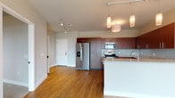 This single-floor unit offers a thoughtfully designed open-concept layout that seamlessly connects the kitchen and living areas. The kitchen is a standout feature, showcasing rich dark-wood cabinetry extending to the ceiling, granite countertops, a light blue glass tile backsplash, and a full suite of stainless steel appliances including a French door refrigerator, electric range, and over-the-range microwave. A peninsula with a built-in sink and pendant lighting overhead adds both functionality and style. Warm hardwood flooring flows throughout the main living areas, complemented by track lighting along the ceiling. The living area is bathed in natural light through expansive floor-to-ceiling sliding glass doors that open to sweeping elevated views of the surrounding cityscape and waterway, creating a bright and airy atmosphere. The bedroom is carpeted for comfort, features a ceiling fan, and offers access to a generously sized walk-in closet with wire shelving systems for ample organization. The bathroom is finished with large-format tile flooring, a wood vanity with granite countertop, a wide mirror with above-vanity lighting, and polished chrome fixtures throughout. The space combines modern finishes with a clean, neutral palette to create a cohesive and comfortable living environment.