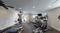 This fitness center is a comprehensively outfitted single-level space designed to support a full range of workout routines. The room features textured ceilings with recessed lighting and a ceiling fan that ensures comfortable air circulation throughout exercise sessions. Neutral-toned walls and dark patterned carpet flooring give the space a clean, functional aesthetic.

The main workout area houses multiple treadmills, an elliptical trainer, and a multi-station cable weight machine, providing options for both cardiovascular and strength training. A wall-mounted flat-screen television is positioned for easy viewing during cardio workouts. A dedicated free-weight station includes a tiered dumbbell rack stocked with a range of weights, alongside a weight bench for versatile strength exercises.

Natural light filters in through several windows fitted with horizontal blinds, offering views to the exterior. A secondary area toward the rear of the space includes a dual water fountain station for hydration, posted fitness center rules, a fire safety station, and restroom access, ensuring the facility meets practical safety and comfort standards. The overall layout is open and organized, allowing users to move freely between equipment.