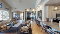 Located in Lake Forest, California, this single-story community clubhouse at 26371 Paloma offers a well-appointed gathering space designed with both functionality and style in mind. The main lounge area is anchored by soaring vaulted ceilings and a dramatic wall of large windows and arched transoms that flood the interior with natural light while framing views of the outdoor pool and pergola. Light wood-tone flooring runs throughout, complementing the warm, neutral color palette. A centerpiece billiards table provides a focal point for the game area, surrounded by stylish lounge seating, accent chairs, and a marble-top coffee table atop a patterned area rug. A wall-mounted flat-screen TV and a decorative tile-accented fireplace add to the entertainment appeal. The adjacent kitchen features a generous quartz island with built-in bar seating, stainless steel appliances, white cabinetry, and elegant pendant lighting. A dedicated business office with glass sliding doors offers a quiet, professional workspace. A pet-friendly station near the entrance provides supplies and resources for four-legged residents. Modern globe and cone pendant fixtures throughout lend a contemporary sophistication, making this clubhouse a versatile and inviting community hub.