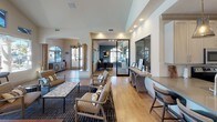 Located in Lake Forest, California, this single-story community clubhouse at 26371 Paloma offers a well-appointed gathering space designed with both functionality and style in mind. The main lounge area is anchored by soaring vaulted ceilings and a dramatic wall of large windows and arched transoms that flood the interior with natural light while framing views of the outdoor pool and pergola. Light wood-tone flooring runs throughout, complementing the warm, neutral color palette. A centerpiece billiards table provides a focal point for the game area, surrounded by stylish lounge seating, accent chairs, and a marble-top coffee table atop a patterned area rug. A wall-mounted flat-screen TV and a decorative tile-accented fireplace add to the entertainment appeal. The adjacent kitchen features a generous quartz island with built-in bar seating, stainless steel appliances, white cabinetry, and elegant pendant lighting. A dedicated business office with glass sliding doors offers a quiet, professional workspace. A pet-friendly station near the entrance provides supplies and resources for four-legged residents. Modern globe and cone pendant fixtures throughout lend a contemporary sophistication, making this clubhouse a versatile and inviting community hub.