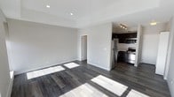 This single-floor space showcases a clean, contemporary design with an open-concept layout that seamlessly connects the living area and kitchen. The dark hardwood flooring runs throughout, adding warmth and contrast against the bright, neutral grey walls and white ceilings. Recessed lighting is installed throughout the living area, providing even, functional illumination, while a track lighting fixture above the kitchen adds focused task lighting.

The kitchen is positioned along one wall, partially separated by a structural divider that lends a sense of definition without closing off the space. It is equipped with a full suite of stainless steel appliances, including a refrigerator, gas range, and microwave, all complemented by rich, dark cabinetry that adds depth and a modern aesthetic. The layout is efficient and well-organized, offering practical workspace for cooking and meal preparation.

The open living area is generously proportioned, benefiting from natural light that streams in and casts bright patches across the flooring. A doorway visible from the main area leads further into the unit, suggesting additional rooms beyond. The overall ambiance is modern, clean, and move-in ready, with a neutral palette that offers flexibility for a variety of interior design styles.