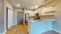 This single-floor residence showcases a thoughtfully designed open-concept layout with a cohesive modern aesthetic throughout. The kitchen serves as a focal point, featuring crisp white shaker-style cabinetry extending to the ceiling, a full suite of stainless steel appliances including a French door refrigerator, electric range, and built-in microwave. A standout teal-accented peninsula with light quartz countertops provides additional workspace and casual seating, while pendant lighting with clear glass fixtures adds a contemporary touch overhead. Warm wood-look luxury vinyl plank flooring flows continuously through the kitchen and living areas, complementing the neutral greige wall tones.

The open living area is generously proportioned, benefiting from natural light through a frosted glass entry door and offering a flexible layout. A dedicated bedroom features plush carpeting and a large picture window with views of a tree-lined streetscape, providing an airy, bright atmosphere.

The bathroom is well-equipped with a wide floating white vanity offering substantial drawer and cabinet storage, a tall linen tower, quartz countertop, and a large mirror. Brushed nickel hardware provides a polished finish. Recessed lighting and a keypad entry door round out the modern, functional details of this well-appointed space.