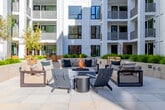 Located at 1350 S Bascom Ave in San Jose, California, this expansive outdoor amenity courtyard is thoughtfully designed to serve as a communal gathering space within a contemporary residential community. The space is defined by two distinct surface zones: a warm hardwood deck and a large-format light stone tile plaza, each creating its own atmosphere while flowing seamlessly together. The hardwood deck area features a prominent central fireplace structure with built-in bench seating, complemented by modern Adirondack-style chairs and side tables — ideal for relaxed social gatherings. Adjacent landscaped planters overflow with ornamental grasses and young olive trees, providing lush greenery that softens the architectural lines. The stone tile plaza extends generously, offering multiple outdoor dining and lounge configurations with contemporary furniture, including curved concrete benches and round pedestal tables. Raised planter beds with mixed grasses and shrubs frame the perimeter, adding texture and privacy. The courtyard is surrounded on multiple sides by the multi-story residential buildings, whose white paneled facades and glass-railed balconies define the horizon. The open-air design maximizes natural light and captures the abundant California sunshine, creating a bright, inviting environment throughout the day.