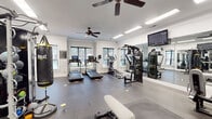 Fitness Center West in Memphis, Tennessee offers a comprehensive workout facility designed to support a wide range of fitness goals. The space is housed within a clean, modern single-floor layout featuring white walls, crown molding, and durable rubberized flooring throughout. Large windows line the walls, flooding the interior with natural light and providing views of the outdoor pool area, creating an energizing atmosphere for exercise.

The facility is equipped with multiple treadmills featuring integrated digital consoles, an elliptical trainer, and a multi-station cable machine for strength training. A full dumbbell rack offers a complete range of free weights, complemented by a flat weight bench for versatile lifting. Functional training is well-supported with a heavy Everlast punching bag, kettlebells, medicine balls, resistance bands, and a BOSU balance trainer. A multi-functional cable and pulley rig rounds out the strength and conditioning options.

Full-length mirrors are mounted along one wall, enhancing the sense of space and allowing users to monitor their form. Ceiling fans are installed throughout to maintain air circulation during workouts, and recessed fluorescent lighting ensures the space is well-lit at all times. A wall-mounted flat-screen television adds entertainment for cardio sessions. Fitness center rules are posted, and a water station is conveniently available for guests.