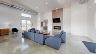 This single-floor space is designed as a contemporary communal amenity facility, combining open-plan living and entertainment zones within a clean, modern aesthetic. Throughout, polished concrete floors and crisp white walls establish a cohesive industrial-modern character, while soaring ceilings with exposed ductwork add architectural interest and a sense of expansive volume.

The main lounge area is anchored by a striking floor-to-ceiling linear fireplace clad in light wood slat paneling, complemented by a wall-mounted television and generous sectional seating in deep blue upholstery. Recessed lighting and large glass entry doors flood the space with natural light, offering views to the exterior.

The kitchen is a standout feature, showcasing an oversized granite-topped island with bar seating, warm wood cabinetry, a gas cooktop, and a stainless steel range hood. Clerestory windows above the perimeter counters maximize daylight throughout this functional space.

A dedicated dining area with wooden tables and upholstered chairs sits adjacent to the kitchen, while a separate game room provides a recreational retreat complete with a ping-pong table and polished concrete floors. An additional flexible open room offers blank-canvas potential for varied uses.
