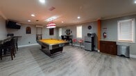 Located at 2930 SW 23rd St in Gainesville, FL, this single-floor entertainment space is a dedicated game room and lounge designed with an unmistakable Florida Gators theme. Bold orange accent walls, a Gators helmet decal, and sports memorabilia throughout establish a spirited atmosphere from the moment you enter.

The space is divided into two primary zones. The game room area features a full-size Murray Professional Series pool table with a striking orange felt surface, illuminated by a classic Tiffany-style hanging pendant light. Adjacent to the pool table is a built-in bar-height seating area, a compact refrigerator, a microwave, and a countertop kitchenette — ideal for entertaining guests.

The lounge area offers generous seating with dark leather sofas and armchairs arranged around a low coffee table, all centered on a large wall-mounted flat-screen television set against a vibrant orange feature wall. Ceiling fans provide comfort throughout, while recessed lighting keeps the space well-lit. Light wood-style flooring runs continuously throughout, lending a clean, modern contrast to the bold color scheme.

Natural light filters in through multiple windows fitted with white blinds. The open floor plan allows both areas to function together seamlessly, making this space well-suited for gatherings, watch parties, and recreational activities.