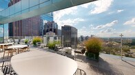 This sophisticated rooftop terrace occupies an elevated position above a bustling urban core, offering unobstructed panoramic views of a dynamic city skyline. The terrace's design is defined by its contemporary architecture, featuring a sleek glass-panel curtain wall facade that reflects the surrounding cityscape and sky, seamlessly blending the built environment with the outdoors.

The terrace is organized into several distinct functional zones, each thoughtfully furnished and landscaped. A dedicated lounge area provides plush, neutral-toned seating arrangements—including sofas and armchairs with warm wood frames—ideal for relaxed gatherings. Nearby, multiple dining areas feature round tables paired with modern wire-frame and teak chairs, suitable for both casual and more formal outdoor dining.

An outdoor kitchen zone is equipped with stainless steel grills and a prep counter, making the space well-suited for entertaining. Throughout the terrace, large planters filled with ornamental grasses, trailing vines, and lush greenery provide natural privacy screening while softening the hard architectural lines.

Large-format stone tiles pave the entire terrace, lending a refined, cohesive aesthetic underfoot. Glass railings along the perimeter ensure uninterrupted sightlines across the skyline. The terrace benefits from abundant natural light and open sky exposure, creating a vibrant and inviting outdoor environment.