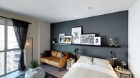 This single-floor space showcases a thoughtfully designed open layout that combines living and sleeping areas into one cohesive environment. The most striking architectural feature is a deep charcoal accent wall that anchors the room, creating a bold visual contrast against the bright white ceiling and lighter surrounding walls. A built-in picture ledge runs horizontally across the accent wall, displaying a curated collection of framed artwork in varying sizes, adding a gallery-like sophistication to the space.

The sleeping area features a neatly made bed with neutral-toned bedding, positioned directly against the accent wall for a grounded, intentional feel. Adjacent to the bed, a compact tan leather sofa introduces a warm, earthy tone that complements the darker backdrop, effectively defining a separate lounge zone within the open plan. A geometric side table with metallic accents serves as a stylish functional element between the two areas.

The flooring consists of light wood-toned planks, contributing to an airy, modern aesthetic. Layered lighting, including a floor lamp and a wall-mounted reading light, enhances the room's ambiance. Greenery near the window adds a natural, refreshing touch to the otherwise urban, contemporary design.