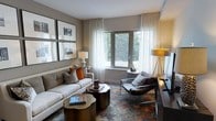 This thoughtfully designed single-floor residence exudes a refined, contemporary aesthetic. The living area serves as the heart of the space, showcasing a curated blend of modern and transitional design elements. A plush white sofa anchors the room, accented with warm burnt-orange and neutral throw pillows that add a lively pop of color against the soft gray walls. A vibrant, multi-toned area rug grounds the seating arrangement, complementing the dark-finished coffee tables that add depth and contrast. A sleek leather accent chair and a gold tripod floor lamp contribute to the room's sophisticated layering of textures and finishes. Large windows dressed in sheer white curtains allow an abundance of natural light to fill the space while framing tranquil views of lush, mature greenery outdoors. A gallery wall featuring framed black-and-white artwork adds a polished, curated touch to the interior. The neutral color palette, warm metallic accents, and thoughtful furniture placement create an inviting yet refined ambiance, making this space ideal for both relaxed everyday living and stylish entertaining.
