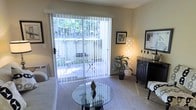 This single-story residence offers a well-organized layout that balances comfort and functionality throughout its living spaces. The living room serves as the heart of the home, featuring neutral-toned walls and soft carpeting that contribute to a warm, inviting atmosphere. A large sliding glass door floods the space with natural light and provides direct access to a private patio, where mature trees and greenery create a tranquil outdoor setting. Decorative touches such as a circular sunburst mirror, framed wall art, and layered accent pillows add personality to the room, while a glass-top coffee table keeps the space feeling open.

Adjacent to the living area, a compact kitchen is thoughtfully appointed with light wood cabinetry, a white range, and under-cabinet lighting. A peninsula counter extends outward to form a breakfast bar with seating for multiple guests, making it a practical space for casual dining. Framed artwork lines the wall along the bar area, adding visual interest to the transition zone between the kitchen and living room. Overall, the space presents a cohesive, comfortable environment well-suited for everyday living.