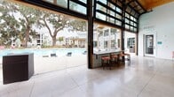 Located at 3 Woodcock Rd in Savannah, Georgia, this single-floor community clubhouse is designed as a dynamic, open-plan gathering space that seamlessly blends indoor comfort with outdoor living. The interior is anchored by soaring ceilings with exposed wooden beams and warm wood-paneled accents, creating a refined yet relaxed atmosphere throughout.

At the heart of the space, a large communal table with ample seating serves as a central hub for socializing and events. Adjacent to it, a well-appointed kitchenette features sleek white cabinetry, a stainless steel sink, a beverage refrigerator, and generous counter space, making the area ideal for hosting. A wall-mounted flat-screen television above the counter adds a functional entertainment element.

The lounge area is furnished with modern seating, including leather sofas, accent chairs, and wall-mounted flat-screen televisions, providing a comfortable space for relaxation and recreation. A game table further enhances the entertainment offerings.

Perhaps the most striking feature is the expansive glass garage-door wall that opens entirely to reveal a resort-style outdoor pool, framed by mature Spanish moss-draped trees — a hallmark of Savannah's iconic landscape. A bar-height counter with red stools straddles the indoor-outdoor boundary, maximizing poolside views. Large format tile flooring, abundant natural light, and clean white walls complete the bright, contemporary aesthetic of this inviting communal space.