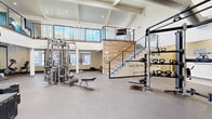 Located at 17249 N 7th St in Phoenix, AZ, this two-level fitness facility offers a generously sized and thoughtfully designed exercise space across two floors. The expansive main level features soaring vaulted ceilings with exposed wood beams and a white shiplap ceiling, lending an open and airy ambiance. Visible industrial-style ductwork adds a modern architectural touch overhead, while recessed lighting ensures a bright and inviting environment throughout.

The main floor is comprehensively equipped for a full-body workout, featuring a row of treadmills, elliptical machines, and stationary bikes for cardio training, alongside a multi-station cable machine, a punching bag, kettlebell racks, weight benches, and plyo boxes for strength and functional training. Motivational quotes are displayed along the walls, contributing to an energizing atmosphere. Durable rubber and hard flooring surfaces are used throughout, appropriate for high-traffic fitness use.

A striking staircase with a reclaimed wood accent wall and sleek metal railings connects the main floor to the upper mezzanine level. This secondary space provides additional workout area with stability balls, a yoga mat, and supplementary equipment, with an open railing offering views down to the main floor below. Wall-mounted televisions are positioned throughout for added convenience.