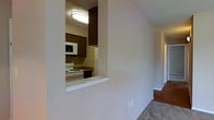 This single-floor residential space offers a practical and functional layout designed for comfortable everyday living. The kitchen is a standout feature, characterized by rich dark wood cabinetry that provides ample storage, a white electric range, and a built-in microwave. A distinctive pass-through window connects the kitchen to the adjacent living area, creating an open sense of communication between the two spaces while maintaining a defined separation. The pass-through ledge doubles as a convenient serving or casual dining surface. The flooring transitions from hard surface material in the kitchen and hallway areas to carpet in the living space, adding warmth and comfort underfoot. Neutral, light-toned walls throughout create a clean and versatile backdrop that allows for easy personalization. A hallway extends toward additional rooms, suggesting a well-organized floor plan with private spaces set apart from the main living area. Overhead flush-mount lighting provides consistent illumination throughout. Overall, the space presents a straightforward, move-in-ready environment with classic design elements suited to a range of lifestyles.