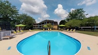 Located at 2255 Country Corners Dr in Columbus, Ohio, this single-level outdoor amenity space centers around a generously sized swimming pool with clear, blue water. The pool area is designed for relaxation and leisure, surrounded by a wide concrete deck that accommodates multiple lounge chairs and side tables, providing ample space for sunbathing and outdoor recreation. Bright green patio umbrellas are positioned throughout the deck, offering shaded seating options for guests throughout the day. The surrounding landscape features mature trees that frame the space and provide a natural, park-like atmosphere. In the background, the multi-story building with bold vertical columns and a distinctive dark roof is visible, lending an architectural backdrop to the outdoor setting. The overall ambiance is open and welcoming, with a well-maintained grounds that contribute to a comfortable and inviting outdoor experience. The combination of lush greenery, structured seating arrangements, and the expansive pool makes this space a prominent recreational feature of the property.