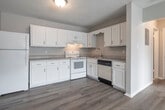 This single-floor unit offers a clean, functional layout with consistent light wood-style flooring running throughout all rooms, creating a cohesive and bright aesthetic. The kitchen is a standout feature, equipped with ample white cabinetry — both upper and lower — providing generous storage space. It includes a full suite of white appliances: a refrigerator, electric range, and dishwasher, complemented by a double-basin sink and light-toned countertops. A flush-mount ceiling fixture provides warm overhead lighting. The open-concept design connects the kitchen seamlessly to the adjacent living area, which is accessed through a paneled interior door. The living spaces are bright and versatile, featuring neutral warm-gray walls, textured ceilings, and large windows with horizontal blinds that allow natural light to fill the rooms. Wall-unit air conditioners are present in the windowed rooms, offering climate control. The bathroom features a full tub-shower combination with white square tile surround and a grab bar. Throughout the unit, clean white baseboards and trim provide a polished finish. The overall ambiance is neutral, tidy, and move-in ready, suited for a variety of living arrangements.