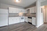 This single-floor unit offers a clean, functional layout with consistent light wood-style flooring running throughout all rooms, creating a cohesive and bright aesthetic. The kitchen is a standout feature, equipped with ample white cabinetry — both upper and lower — providing generous storage space. It includes a full suite of white appliances: a refrigerator, electric range, and dishwasher, complemented by a double-basin sink and light-toned countertops. A flush-mount ceiling fixture provides warm overhead lighting. The open-concept design connects the kitchen seamlessly to the adjacent living area, which is accessed through a paneled interior door. The living spaces are bright and versatile, featuring neutral warm-gray walls, textured ceilings, and large windows with horizontal blinds that allow natural light to fill the rooms. Wall-unit air conditioners are present in the windowed rooms, offering climate control. The bathroom features a full tub-shower combination with white square tile surround and a grab bar. Throughout the unit, clean white baseboards and trim provide a polished finish. The overall ambiance is neutral, tidy, and move-in ready, suited for a variety of living arrangements.