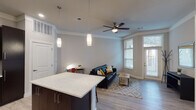 This single-floor residence offers a thoughtfully designed open-concept layout that seamlessly connects the kitchen and living areas. The kitchen is a standout feature, equipped with rich dark cabinetry, stainless steel appliances including a refrigerator, range, microwave, and dishwasher, complemented by a large granite-topped island that provides ample prep and dining space. Pendant lighting illuminates the kitchen and transitions naturally into the living area, where light wood-look flooring, crown molding, and a ceiling fan contribute to a clean, modern aesthetic. Large windows and a glass-panel entry door flood the living space with natural light, creating an open and airy atmosphere. The bedroom is carpeted for comfort and features a sizable window with exterior views, offering a quiet and restful retreat. The bathroom includes a full tub and shower combination with built-in shelving and a curved shower rod. A dedicated in-unit laundry area adds everyday convenience. Neutral tones throughout the space provide a versatile backdrop, while the combination of recessed and pendant lighting ensures a well-lit environment at all hours. The overall design balances functionality with contemporary style.