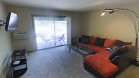 This single-floor space centers around a comfortable living room finished with neutral-toned carpet that extends throughout. The room is anchored by a large L-shaped sectional sofa upholstered in a bold rust-orange fabric and accented with dark decorative pillows, paired with a sleek glass-top coffee table supported by a metal frame. A wall-mounted light fixture provides ambient illumination, complementing the natural light that filters generously into the room. The most prominent architectural feature is the wide sliding glass door, framed by sheer white curtains, which opens directly onto a private balcony. The balcony is furnished with outdoor seating and a small dining table and chairs, offering a relaxed outdoor extension of the living space. Through the glass, lush greenery and open surroundings are visible, enhancing the sense of connection to the outdoors. The overall ambiance is casual and functional, with a neutral color palette on the walls and flooring providing a versatile backdrop that allows the bold furnishings and outdoor views to stand out as focal points.