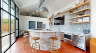 This single-floor space combines industrial and contemporary design elements to create a dynamic, open communal environment. The architecture is immediately striking, with soaring vaulted ceilings that rise to a peaked roofline, exposed ductwork, and oversized black-framed, floor-to-ceiling windows that flood the interior with natural light and provide views of the surrounding outdoor courtyard and greenery.

The centerpiece of the space is a curved bar constructed from exposed brick, topped with a smooth white quartz countertop and surrounded by adjustable gold-toned bar stools with wooden seats. Behind the bar, a decorative terrazzo-style accent wall serves as the backdrop for open wooden shelving and a wall-mounted flat-screen television, with stainless steel appliances including a microwave and beverage refrigerator integrated beneath. A bold industrial pendant light hangs overhead, complementing the visible HVAC ductwork above.

The lounge area features plush orange barrel chairs arranged around paired circular coffee tables, contributing warmth and a pop of color against the polished terracotta-toned concrete floors. A wall-mounted screen and additional seating with compact café-style tables offer flexible gathering options. The direct access to the outdoor patio further enhances the open, connected ambiance of the space.