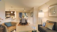 This single-floor residence is designed with an open-concept layout that allows the living, dining, and kitchen areas to flow naturally into one another. The interior is finished in warm, neutral tones, with light beige walls and large-format tile flooring that enhances the sense of spaciousness and continuity throughout. The dining area serves as a central focal point, anchored by a dark rectangular table surrounded by upholstered grey chairs and accented by a bold, colorful abstract artwork that adds a vibrant contrast to the soft palette. A warm-toned area rug beneath the dining set adds texture and definition to the space. Recessed and track lighting provides targeted illumination, while a stylish pendant light near the kitchen adds a decorative touch. The living area, partially visible in the foreground, features a charcoal sofa accented with blue patterned cushions and a delicate orchid centerpiece, contributing to an elegant, curated ambiance. A tall, decorative floor lamp with a chrome base anchors the transition between the living and dining spaces. The overall atmosphere is polished, bright, and inviting, with thoughtful design details throughout.