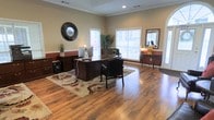 This single-floor professional office space combines functionality with a warm, inviting aesthetic. The main office area is anchored by rich hardwood flooring that extends throughout, lending a sense of warmth and sophistication to the environment. A substantial executive desk with a leather chair serves as the focal point of the workspace, complemented by a decorative area rug that adds texture and definition to the room.

Crown molding frames the ceiling, adding a refined architectural detail that elevates the overall design. Multiple windows fitted with white plantation shutters allow generous natural light to fill the space, creating a bright and welcoming atmosphere. The neutral wall tones, accented by a warm wainscoting detail along the lower walls, contribute to a professional yet comfortable ambiance.

Decorative touches throughout the space include a round ornate wall mirror, a tall indoor plant, and an elegant sideboard with a classic lamp and mirror — all of which enhance the room's polished character. Recessed lighting provides additional illumination, ensuring the space remains well-lit throughout the day. Overall, this office presents a well-appointed, professional environment that balances practical workspace needs with tasteful interior design.