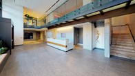 Located in Chicago's vibrant West Loop neighborhood, this single-floor commercial lobby at 147–169 N Desplaines St presents a sophisticated and thoughtfully designed arrival experience. The entrance is framed by dramatic floor-to-ceiling glass curtain walls, flanked by built-in planter beds featuring lush greenery that soften the industrial-modern aesthetic. Inside, the expansive lobby is anchored by a contemporary white reception desk with warm wood accents, set beneath an open mezzanine level outfitted with glass-panel railings and exposed structural beams that add architectural depth. Stone-tiled flooring flows throughout, lending a polished, durable foundation to the space. A standout lounge area features a sleek, freestanding linear fireplace set against a wall of steel-framed windows offering views of tree-lined streetscapes. A large-scale, vibrant abstract mural in warm amber and golden tones serves as a bold artistic focal point, complemented by plush seating. A secondary corridor leads to a bank of stainless steel elevators, where herringbone wood flooring, a built-in service counter, and curated wall-mounted displays create a refined transition space. Throughout, the interplay of natural light, contemporary art, and high-quality materials establishes a welcoming yet impressive professional environment.