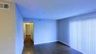 This single-floor residence offers a clean, straightforward layout designed for comfortable everyday living. The main living area is immediately welcoming, featuring dark wood-style flooring that contrasts crisply with soft blue-painted walls and bright white baseboards. A textured ceiling adds subtle character to the space, while a ceiling-mounted HVAC vent ensures year-round climate comfort. Natural light filters into the room through a large window fitted with vertical blinds, providing both brightness and privacy control. A hallway leads from the main living area deeper into the home, hinting at additional rooms beyond — illuminated by a ceiling light fixture that keeps the passage well-lit. The overall design is minimal and neutral, offering a versatile canvas well-suited to a variety of furnishing styles. The consistent flooring and wall tones throughout create a cohesive, unified atmosphere across the space. The layout feels open and uncluttered, making efficient use of the available footprint on the single floor. Clean lines, practical finishes, and thoughtful lighting combine to give this space a functional yet inviting character.