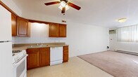 Located at 24350 Lake Shore Blvd in Euclid, Ohio, this single-floor unit offers a practical and straightforward layout designed for efficient use of space. The kitchen serves as a central functional hub, equipped with warm-toned oak cabinetry featuring arched door detailing, a white gas range, over-the-range microwave, refrigerator, dishwasher, and a double basin sink. White subway tile backsplash lines the walls, complementing the neutral countertops and light vinyl flooring underfoot. A ceiling fan overhead provides added air circulation to the open kitchen and living area, which transitions seamlessly from vinyl to carpet. The living space is bright and open, with vertical blinds filtering natural light across the carpeted floors, and a baseboard heating system along the perimeter wall ensures warmth throughout. The bathroom is finished with classic white square tile extending along the walls, a white vanity with an inset medicine cabinet mirror, and a clean, minimalist aesthetic throughout. A hallway connects the rooms and contributes to the overall flow of the unit. The space presents a clean, functional interior well-suited for straightforward, comfortable living.