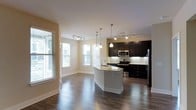 This single-floor space showcases an open-concept layout that integrates the kitchen and living areas into a cohesive, functional environment. The kitchen is a standout feature, outfitted with rich, dark espresso cabinetry that extends to the upper and lower sections of the walls, creating a bold contrast against the light cream-colored walls and white countertops. A central island with a granite countertop serves as both a workspace and a casual gathering point, accented by elegant pendant lighting that hangs overhead. Stainless steel appliances, including a built-in microwave, range, and refrigerator, add a polished, contemporary finish. Track lighting is installed throughout both the kitchen and the adjoining living area, ensuring consistent and versatile illumination. The living area is characterized by its generous proportions and dark hardwood flooring, which flows seamlessly throughout the entire space. Multiple large windows fitted with white horizontal blinds allow abundant natural light to fill the interior, creating a bright and welcoming ambiance. The neutral wall tones complement the overall design, lending the space a clean, modern aesthetic well-suited for everyday living.