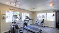 This fitness center is designed to support an active lifestyle, offering a dedicated space for cardiovascular training and exercise. The room is outfitted with a variety of cardio equipment, including treadmills, an elliptical trainer, and a recumbent bike, providing options for users of all fitness levels. Natural light fills the space through multiple windows, which also offer views of the surrounding outdoor landscape, including palm trees and open-air corridors that contribute to an inviting atmosphere. Motivational phrases such as "Every Day Is a Good Day to Work Out" and "No Pain, No Gain" are displayed along the walls, adding an energizing touch to the environment. The flooring features a durable, textured carpet suitable for a fitness setting, while track lighting along the ceiling ensures the space is well-lit throughout the day. The textured ceiling and neutral wall tones keep the aesthetic clean and functional. Overall, the fitness center is bright, practical, and thoughtfully arranged to encourage regular use and an active lifestyle.