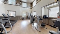 Located at 16601 Foothill Blvd, this well-appointed fitness center occupies a single floor and offers a comprehensive workout environment designed to accommodate a wide range of exercise needs. The facility boasts soaring double-height ceilings that create an open, airy atmosphere, complemented by large windows fitted with blinds that allow abundant natural light to fill the space. Motivational fitness-themed typography runs along the upper walls, adding an energetic visual element throughout.

The workout floor is finished with smooth, light-toned hardwood-style flooring, providing a clean and durable surface for all types of exercise. The cardio zone features a row of Matrix elliptical trainers, each equipped with built-in digital displays, positioned in front of wall-mounted flat-screen televisions for an engaging workout experience. Cycling bikes and additional cardio equipment round out this section.

The strength training area includes a multi-station cable and weight machine setup alongside benches, catering to a full range of resistance exercises. A dedicated free weights section offers a tiered dumbbell rack with a full set of weights, accompanied by floor-to-ceiling mirrors that maximize visual space and support proper form. Built-in shelving units neatly store stability balls, resistance accessories, and yoga mats. A television and cabinetry unit provides added convenience within the space.
