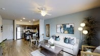 This single-floor residence showcases an open-concept design that seamlessly connects the living, dining, and kitchen areas into one cohesive, functional space. The living area is anchored by a white sofa accented with teal and patterned throw pillows, complemented by a wood-finish coffee table and a soft area rug. A ceiling fan with integrated lighting fixture is centrally mounted overhead, enhancing airflow and comfort. The walls are painted in a soothing sage-green tone, lending the space a calm, contemporary ambiance, further accented by a large abstract canvas artwork.

Adjacent to the living room, the dining area accommodates a glass-top table with modern upholstered chairs, offering a clean and stylish setting for meals. A framed mirror nearby adds depth and light to the space.

The kitchen features dark cabinetry contrasting with light countertops, stainless steel appliances including a refrigerator and microwave, and recessed lighting. Wood-style flooring flows consistently throughout all areas, tying the open-plan layout together cohesively. Recessed ceiling lights supplement natural light, creating a bright and welcoming atmosphere throughout.