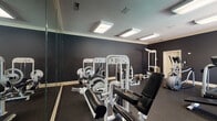 Located at 2140 Pine Tree Ln in Rocky Mount, North Carolina, this fitness center offers a well-appointed, single-floor workout facility designed to support a full range of exercise routines. The space is finished with dark, charcoal-toned walls that lend a modern and focused atmosphere, complemented by crown molding and a textured ceiling fitted with bright flush-mount panel lighting that ensures consistent, even illumination throughout.

The strength training area is generously stocked with a variety of white cable and resistance machines, including leg press, lat pulldown, and cable crossover stations, along with a dedicated dumbbell rack and a flat workout bench. Full-length mirrors line one wall, enhancing the sense of space and allowing users to monitor their form during exercise.

The cardio zone is equipped with at least one treadmill and an elliptical trainer, positioned near large windows that provide natural light and views of an outdoor pool and surrounding greenery, creating an inviting and energizing workout environment. Durable rubber flooring covers the entire space, providing both safety and comfort underfoot.

A wall-mounted clock and safety signage add practical, functional touches to the room. The overall layout is thoughtfully organized, offering ample space to move between equipment and accommodating both strength and cardio training needs in a clean, professional setting.