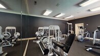 Located at 2140 Pine Tree Ln in Rocky Mount, North Carolina, this fitness center offers a well-appointed, single-floor workout facility designed to support a full range of exercise routines. The space is finished with dark, charcoal-toned walls that lend a modern and focused atmosphere, complemented by crown molding and a textured ceiling fitted with bright flush-mount panel lighting that ensures consistent, even illumination throughout.

The strength training area is generously stocked with a variety of white cable and resistance machines, including leg press, lat pulldown, and cable crossover stations, along with a dedicated dumbbell rack and a flat workout bench. Full-length mirrors line one wall, enhancing the sense of space and allowing users to monitor their form during exercise.

The cardio zone is equipped with at least one treadmill and an elliptical trainer, positioned near large windows that provide natural light and views of an outdoor pool and surrounding greenery, creating an inviting and energizing workout environment. Durable rubber flooring covers the entire space, providing both safety and comfort underfoot.

A wall-mounted clock and safety signage add practical, functional touches to the room. The overall layout is thoughtfully organized, offering ample space to move between equipment and accommodating both strength and cardio training needs in a clean, professional setting.
