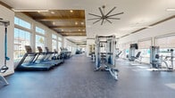 Located at 1600 LPGA Blvd in Daytona Beach, Florida, this well-appointed fitness center offers a bright, open layout designed to support a comprehensive workout experience. The space features soaring ceilings with elegant wood-paneled coffered accents, complemented by large oversized windows that flood the interior with natural light and frame views of the surrounding palm-lined landscape. Durable speckled rubber flooring spans the entire area, providing both functionality and a polished aesthetic throughout.

The facility is comprehensively equipped with a long row of cardio machines — including treadmills, ellipticals, and stair climbers — positioned along the windowed perimeter to take full advantage of the outdoor views. The strength training section offers a multi-station cable machine, a tiered dumbbell rack with a full range of free weights, and multiple adjustable weight benches. Suspension training equipment is also available for functional fitness workouts.

Oversized industrial ceiling fans are strategically placed throughout the space to maintain airflow and comfort. A wall-mounted television provides entertainment, while mirrors along one wall enhance the sense of openness. The entrance features glass French doors flanked by large windows and a decorative chandelier, creating a welcoming and upscale first impression befitting this Daytona Beach location.