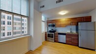 This single-floor urban residence offers a thoughtfully designed open-concept layout that balances modern finishes with functional living. The kitchen serves as a standout feature, showcasing rich warm-toned wood cabinetry, granite countertops, a white tile backsplash, and a full suite of stainless steel appliances including a range, microwave, dishwasher, and refrigerator. Light hardwood flooring extends throughout the main living area, lending warmth and cohesion to the space.

The living area is generously proportioned and bathed in natural light, courtesy of an expansive bank of large windows fitted with horizontal blinds. These oversized windows frame striking views of the surrounding urban skyline, reinforcing the elevated city setting of this residence. The clean white walls and high ceilings enhance the sense of openness throughout.

The bedrooms feature neutral carpeting and their own large windows that continue the urban views, with one room offering a dramatic three-panel window arrangement looking out onto neighboring city architecture. The overall design is contemporary and clean, with a neutral palette that provides a versatile backdrop. The space is well-suited for those seeking a modern urban lifestyle with an abundance of natural light and city character.