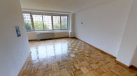This single-floor space is characterized by its clean, minimalist design and well-maintained interior. The standout feature is the classic parquet hardwood flooring, laid in a traditional block pattern that adds warmth and timeless elegance throughout the room. The flooring's polished finish reflects light beautifully, enhancing the overall brightness of the space.

A large picture window dominates one wall, framing views of mature trees and neighboring buildings, while allowing generous amounts of natural light to flood the interior. Beneath the window, a built-in radiator provides heating, a detail consistent with established residential architecture. The walls are finished in clean white, creating a neutral backdrop that complements the warm tones of the hardwood floors.

The space is generously proportioned, offering an open and airy feel that provides flexibility for various furniture arrangements. The room's simple, uncluttered design ensures that its architectural features — particularly the flooring and window — remain the focal points. Overall, the space presents a bright, inviting atmosphere with classic details and urban views.
