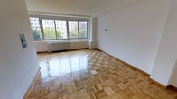 This single-floor space is characterized by its clean, minimalist design and well-maintained interior. The standout feature is the classic parquet hardwood flooring, laid in a traditional block pattern that adds warmth and timeless elegance throughout the room. The flooring's polished finish reflects light beautifully, enhancing the overall brightness of the space.

A large picture window dominates one wall, framing views of mature trees and neighboring buildings, while allowing generous amounts of natural light to flood the interior. Beneath the window, a built-in radiator provides heating, a detail consistent with established residential architecture. The walls are finished in clean white, creating a neutral backdrop that complements the warm tones of the hardwood floors.

The space is generously proportioned, offering an open and airy feel that provides flexibility for various furniture arrangements. The room's simple, uncluttered design ensures that its architectural features — particularly the flooring and window — remain the focal points. Overall, the space presents a bright, inviting atmosphere with classic details and urban views.