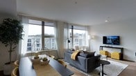 This single-floor residence showcases a thoughtfully designed open-concept living and dining area that emphasizes natural light and modern aesthetics. Large floor-to-ceiling windows span the living room walls, flooding the interior with daylight while offering views of the surrounding residential buildings and mature treetops beyond. Sheer white curtains frame the windows, softening the light and adding an airy quality to the space.

The living area is anchored by a plush, dark grey sectional sofa accented with mustard yellow throw pillows and a cozy blanket, creating a warm contrast against the neutral tones of the room. A flat-screen television is mounted on a sleek media unit to one side, while floor and arc lamps provide ambient lighting options. The space transitions seamlessly into a dining area, where a dark-toned dining table is paired with light wood chairs, all set atop a natural jute-style area rug over rich hardwood flooring.

Decorative geometric lanterns and candle holders serve as stylish centerpieces on the dining table, adding an artistic touch. The clean white ceilings and walls maintain a fresh, contemporary feel throughout, making this a well-appointed and visually cohesive living space.