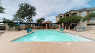 This expansive outdoor pool area offers a well-appointed resort-style retreat designed for relaxation and social gatherings. The centerpiece is a generously sized swimming pool with clear turquoise water, accented by large decorative planters in a matching teal finish placed along the pool deck. Ample lounge chairs with bright blue cushions line the poolside, providing plenty of space for sunbathing and leisure.

Surrounding the pool are multiple covered pergola structures with dark wood beams and stone columns, offering shaded seating areas furnished with wicker and metal tables and chairs. One pavilion features an elegant chandelier and lush climbing greenery overhead, creating a garden-like atmosphere. Another covered area houses a striking floor-to-ceiling stacked-stone fireplace with a terracotta tile roof, ideal for cooler evenings.

An outdoor kitchen station equipped with a stainless steel grill and stone countertops provides convenient space for outdoor cooking and entertaining. Blue market umbrellas are positioned throughout the deck, offering additional shade over casual dining arrangements. The entire area is enclosed by mature trees that provide natural privacy and a serene backdrop, complementing the Mediterranean-influenced architectural details visible throughout the space.