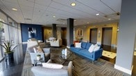 Located at 414 N Forest Park Blvd in Knoxville, Tennessee, this single-floor space presents a thoughtfully designed communal lounge area that combines modern aesthetics with comfortable functionality. The open-plan layout is anchored by a bold blue sofa accented with decorative throw pillows, complemented by an arrangement of neutral-toned armchairs and settees that create distinct yet connected seating zones throughout the space. Structural columns in a dark charcoal finish add architectural definition while subtly delineating areas within the open floor plan. The flooring features patterned carpeting in earthy tones, contributing texture and visual warmth underfoot. Recessed lighting set into a drop-tile ceiling provides consistent, even illumination throughout. The color palette blends deep navy blue accent walls with warm yellow tones, creating a vibrant yet cohesive atmosphere. Glass-top side tables and small potted greenery add refined finishing touches. Framed artwork adorns the walls, enhancing the space's polished, contemporary character. Corridors visible in the background suggest connectivity to additional areas, making this lounge an inviting central hub well-suited for gathering, relaxing, or casual interaction.