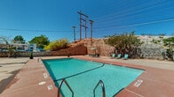 This outdoor pool area offers a well-appointed recreational space designed for relaxation in a desert setting. The centerpiece is a generously sized rectangular swimming pool with clear turquoise water, ranging from a shallow end to a deeper section, accessible via stainless steel entry rails on multiple sides. The pool is edged by a broad, terra-cotta-toned concrete deck that provides ample space for lounging and circulation.

The deck is furnished with teal lounge chairs positioned to take advantage of the open sky and natural surroundings, along with a shaded seating area featuring a green umbrella, table, and chairs — ideal for outdoor dining or relaxation out of direct sun. Potted plants add subtle greenery to the hardscape environment.

The space is bordered by a rugged natural rock formation and stone retaining wall to one side, lending a striking, distinctly southwestern character to the setting. An adobe-style stucco building with arched architectural detailing is visible adjacent to the pool area, reinforcing the regional aesthetic. The surrounding landscape, characterized by arid terrain, desert vegetation, and mountain views in the distance, creates an open, sun-drenched ambiance consistent with the American Southwest.