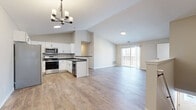 This two-story home offers a well-designed layout across two floors, with the majority of living space situated on the upper level. The entrance leads into a compact lower floor, which transitions via staircase to the expansive main living areas above.

The open-concept kitchen and living room serves as the heart of the home, featuring vaulted ceilings that lend a sense of height and airiness. The kitchen is outfitted with white shaker-style cabinetry, dark countertops, and a full suite of stainless steel appliances including a refrigerator, range, microwave, and dishwasher. Light wood-style flooring extends throughout the main living area, which is illuminated by a chandelier and benefits from a sliding glass door that opens to a balcony with outdoor views.

The four bedrooms are carpeted and receive natural light through windows. The generously sized primary bedroom includes a large walk-in closet with wire shelving systems. A second closet provides additional storage elsewhere on the floor. The two bathrooms feature tub-shower combinations with clean white surrounds and wood-look flooring. Neutral warm-toned walls throughout the home create a cohesive and understated aesthetic.