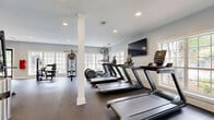 Located at 7150 W Peachtree Dunwoody Rd NE in Atlanta, GA, this fitness facility is thoughtfully divided into two distinct exercise rooms, each designed to support a comprehensive workout experience. The main strength and cardio room features durable rubber flooring, recessed lighting, and large windows that invite natural light and offer views of the surrounding greenery. This area is outfitted with multiple treadmills, an elliptical, a Smith machine with a weight bench, a cable machine system, TRX suspension trainers, and a wall-mounted television — providing a full range of resistance and cardiovascular training options. Floor-to-ceiling mirrors along one wall amplify the sense of space and allow for form monitoring during exercise. The second room offers a bright, open studio environment with warm hardwood floors and expansive multi-pane windows on multiple walls, flooding the space with natural light and scenic outdoor views. This room accommodates cycling bikes with individual monitors, a functional training rack stocked with medicine balls, kettlebells, and stability balls, as well as a weight bench for versatile use. Together, the two rooms form a well-rounded, professionally appointed fitness center suited for a wide variety of training disciplines.