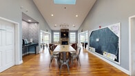 This single-floor multi-purpose space is designed to accommodate a variety of social and recreational activities within a well-appointed, open layout. The main gathering area is anchored by a dramatic floor-to-ceiling stacked stone fireplace with a mounted flat-screen television above a slate mantel, flanked by built-in bench seating with dark granite tops and white wainscoting panels. Natural light streams in through multiple large windows fitted with horizontal blinds.

The vaulted ceiling rises prominently above the central area, enhanced by a skylight that introduces additional daylight, while recessed lighting and track fixtures ensure consistent illumination throughout. Warm-toned hardwood flooring extends across the entire space, contributing to a cohesive and inviting atmosphere.

A central dining area features a large communal wood table surrounded by industrial-style metal chairs. A built-in nook along one wall, accented with stacked stone tile, houses a small beverage or coffee station. A large chalkboard wall serves as a functional and informal focal point nearby.

Adjoining areas include a game room equipped with a pool table and ping pong table, as well as a seating lounge with leather armchairs. French doors and multiple entry points connect the zones seamlessly, creating a flexible, activity-rich environment.