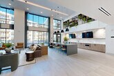 Located at 4015 South Blvd in Charlotte, NC, this contemporary amenity space delivers an impressive blend of modern design and multifunctional living. The single-floor layout spans a generous open footprint, seamlessly connecting several distinct activity zones under soaring double-height ceilings that flood the interior with natural light through dramatic floor-to-ceiling glass walls. The main lounge area showcases a striking marble-clad accent wall with integrated cabinetry, a large central island with bar seating, and pendant lighting above light wood flooring—creating an inviting hub ideal for socializing or working. Plush, thoughtfully arranged seating areas invite relaxed gathering, complemented by lush greenery and curated open shelving above. A statement concrete fireplace wall, flanked by warm wood cladding and built-in shelving, anchors a cozy secondary lounge with ample seating. The game and entertainment wing features exposed wood beam ceiling accents, a billiards table, shuffleboard, and high-top seating with wall-mounted displays. A dedicated open event space with light hardwood flooring, art gallery-style wall displays, and a round dining table rounds out this versatile facility. Throughout, the design balances clean white walls, natural wood tones, and bold architectural accents, creating an elevated, contemporary atmosphere.