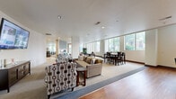 This well-appointed single-floor space offers a generous and open layout encompassing a living room, dining room, and fully equipped kitchen. The interior design blends modern and contemporary elements, with a warm, welcoming ambiance throughout.

The living area features plush seating arrangements, including a neutral-toned sofa and patterned accent chairs set atop area rugs, with a wall-mounted flat-screen television providing a central focal point. Recessed lighting and smooth white ceilings contribute to the bright, airy feel of the space.

The dining area is furnished with dark-wood high-top tables and chairs, positioned alongside large windows that frame lush outdoor greenery and invite abundant natural light into the room. The seamless transition between hardwood flooring and carpeted areas clearly delineates the different functional zones.

The kitchen is a standout feature, showcasing rich dark-wood cabinetry, stainless steel appliances including a French door refrigerator and built-in microwave, light stone countertops, and a spacious island with bar seating for three. Vibrant yellow pendant lights add a striking design accent. A separate lounge area near the entry features bold artwork, a living green wall, and floor-to-ceiling windows that flood the space with natural light.