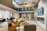 Located at 3616 N Rocky River Road in Monroe, NC, this thoughtfully designed community clubhouse offers a sophisticated and welcoming atmosphere across a single open floor. The living room serves as the centerpiece of the space, boasting dramatically high ceilings adorned with eye-catching circular pendant chandeliers and exposed wood beam accents. A striking living wall with lush greenery adds a natural touch overhead, while large windows usher in ample natural light. The seating area is anchored by a plush sectional sofa in warm amber tones, complemented by mid-century modern accent chairs and layered area rugs that define the lounge zone. A white brick fireplace with a mounted flat-screen TV serves as a focal point, flanked by bold geometric wallpaper and a built-in bench. Large-scale sculptural wall art adds an artistic dimension to the room. The kitchen area features a generous butcher block island bar with seating, dark cabinetry, textured sage-green tile backsplash, and pendant lighting overhead. Adjacent to the kitchen, a billiards table with a brass pendant light above anchors a dedicated game area with its own stylish area rug. Throughout, the design blends earthy greens, warm neutrals, and natural materials to create an inviting, upscale communal environment.